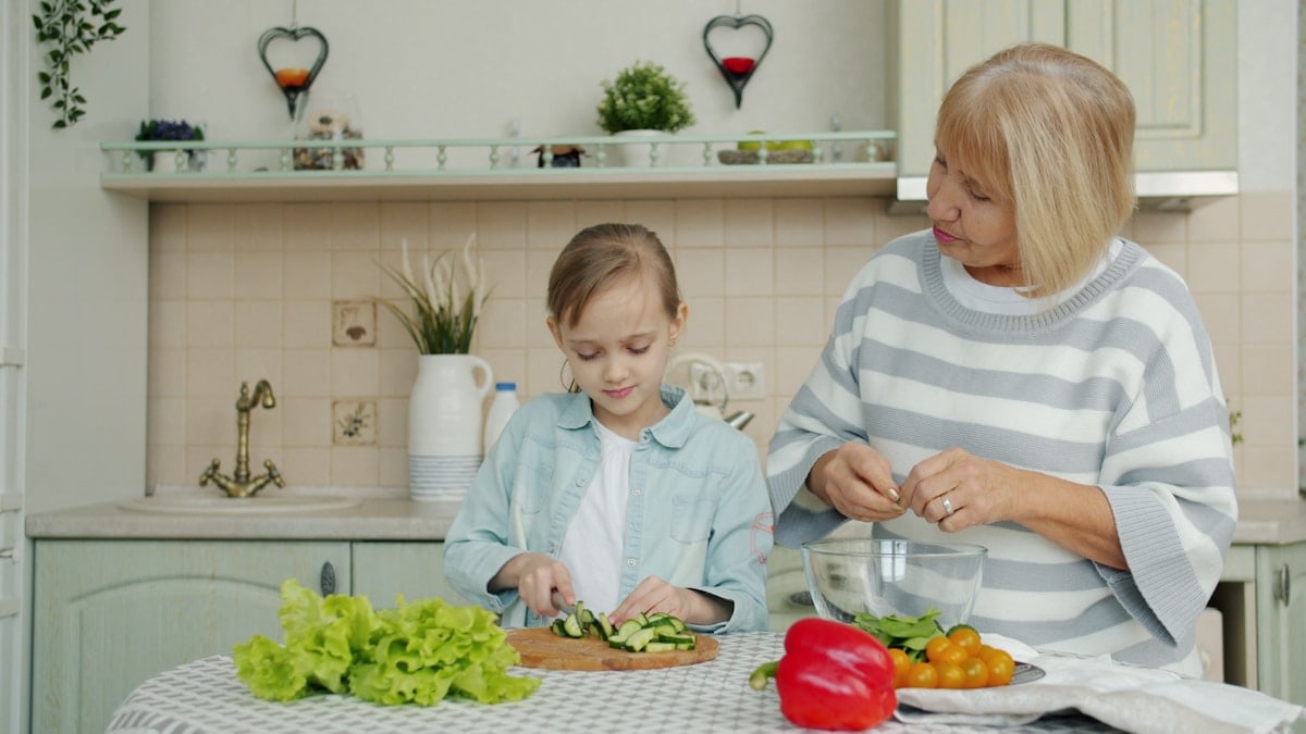 Cute little girl and elderly woman granny are cooking salad at kitchen table together preparing meal and talking. Lifestyle, nutrition and people concept.